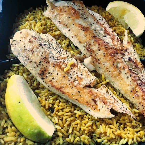 Two grilled fish fillets seasoned with pepper, served over yellow rice with lemon wedges.